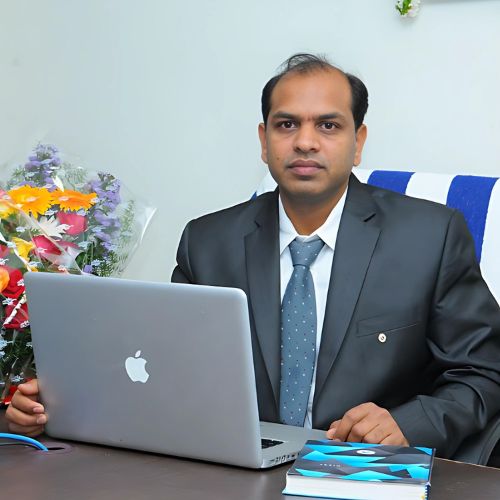 A professional sitting at a desk with a laptop, wearing a dark suit and tie, next to a bouquet of flowers, symbolizing healthcare expertise at Sai Ayush Ayurveda Hospital in KPHB, Hyderabad.